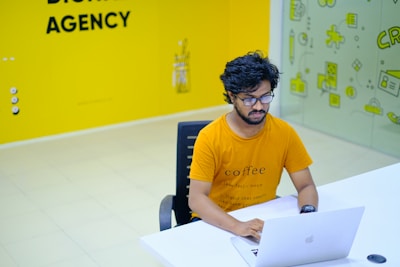 A person with glasses and a beard is sitting at a white desk, working on a laptop. They are wearing an orange t-shirt with text on it. The surroundings include a brightly colored office space with a predominantly yellow theme. There is a wall with the words 'Digital Agency' and a glass partition featuring playful icons.