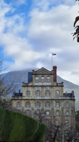 A historic stone building labeled 'Cascade Brewery' with a classical architectural style, featuring multiple windows and a flagpole on the roof. The building is set against a backdrop of a partly cloudy sky and a mountain, with lush greenery and trees in the foreground.
