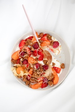 A bowl filled with creamy yogurt topped with vibrant strawberries, raspberries, sliced apricots, and chunks of granola. A pink spoon rests inside the bowl, which is set against a soft white background.