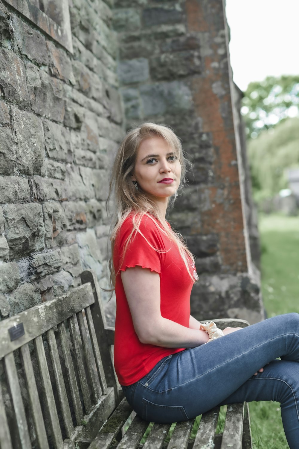 woman in red shirt and blue denim jeans sitting on brown wooden bench