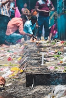People are gathered around a small fire with various offerings placed on the ground, including candles, incense sticks, and flowers. The setting suggests a ceremonial or religious activity with individuals focused on the offerings.