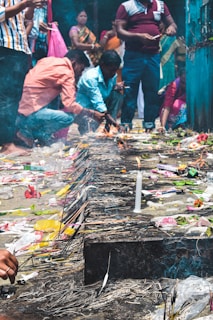 People are gathered around a small fire with various offerings placed on the ground, including candles, incense sticks, and flowers. The setting suggests a ceremonial or religious activity with individuals focused on the offerings.