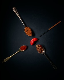 A dark background highlights three vintage spoons filled with different types of spices. One spoon holds ground spice, another has crushed red pepper flakes, and the last contains a fine red powder, possibly paprika. A bright red pepper is positioned between the spoons, and a vanilla pod is also visible.
