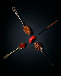 A dark background highlights three vintage spoons filled with different types of spices. One spoon holds ground spice, another has crushed red pepper flakes, and the last contains a fine red powder, possibly paprika. A bright red pepper is positioned between the spoons, and a vanilla pod is also visible.