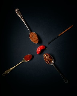 A dark background highlights three vintage spoons filled with different types of spices. One spoon holds ground spice, another has crushed red pepper flakes, and the last contains a fine red powder, possibly paprika. A bright red pepper is positioned between the spoons, and a vanilla pod is also visible.