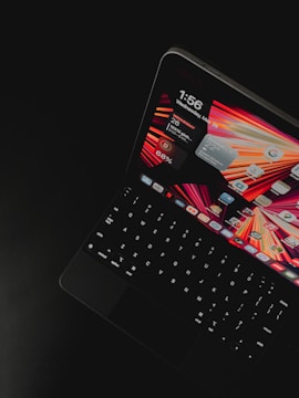 A tablet device with a keyboard attachment is displayed against a dark background. The screen shows a digital layout with a vibrant wallpaper featuring red, orange, and yellow colors. Various application icons and widgets are visible, including a calendar, weather information, and a battery life indicator.