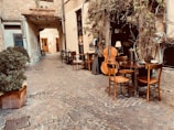 A quaint cobblestone alleyway with an outdoor seating area featuring wooden chairs and tables. A large double bass and a French horn are placed against the wall, suggesting a musical setting. Plants with green leaves climb the walls, adding to the cozy atmosphere.