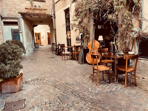 A quaint cobblestone alleyway with an outdoor seating area featuring wooden chairs and tables. A large double bass and a French horn are placed against the wall, suggesting a musical setting. Plants with green leaves climb the walls, adding to the cozy atmosphere.