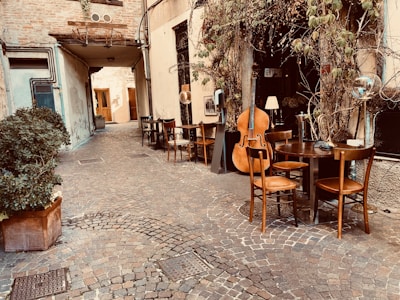 A quaint cobblestone alleyway with an outdoor seating area featuring wooden chairs and tables. A large double bass and a French horn are placed against the wall, suggesting a musical setting. Plants with green leaves climb the walls, adding to the cozy atmosphere.