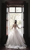 A woman in an elaborate, lace-embroidered wedding dress stands in front of an open window, looking outside. The dress features a full skirt and long sleeves with intricate lace detailing. Sunlight filters through the window, casting a soft glow around her.