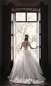 A woman in an elaborate, lace-embroidered wedding dress stands in front of an open window, looking outside. The dress features a full skirt and long sleeves with intricate lace detailing. Sunlight filters through the window, casting a soft glow around her.