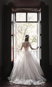 A woman in an elaborate, lace-embroidered wedding dress stands in front of an open window, looking outside. The dress features a full skirt and long sleeves with intricate lace detailing. Sunlight filters through the window, casting a soft glow around her.