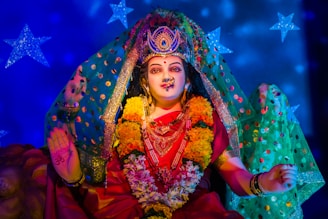 A beautifully adorned Hindu deity statue with intricate jewelry, floral garlands, and a vibrant red saree. The statue has a serene expression, surrounded by a colorful backdrop with blue stars. The hands are posed in symbolic gestures.