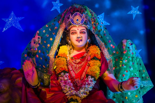 A beautifully adorned Hindu deity statue with intricate jewelry, floral garlands, and a vibrant red saree. The statue has a serene expression, surrounded by a colorful backdrop with blue stars. The hands are posed in symbolic gestures.