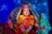 A beautifully adorned Hindu deity statue with intricate jewelry, floral garlands, and a vibrant red saree. The statue has a serene expression, surrounded by a colorful backdrop with blue stars. The hands are posed in symbolic gestures.