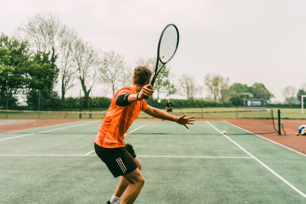 homme en chemise orange et short noir tenant une raquette de tennis noire et blanche