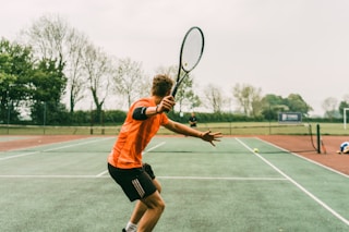 man in orange shirt and black shorts holding black and white tennis racket