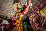 A vibrant and intricately designed statue of a deity, adorned with elaborate jewelry and colorful garments, stands commanding and powerful. A fierce tiger is positioned by the deity's side, showcasing its strength and loyalty. The setting is decorated with floral garlands and a patterned dome, enhancing the sacred and auspicious atmosphere.