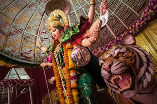 A vibrant and intricately designed statue of a deity, adorned with elaborate jewelry and colorful garments, stands commanding and powerful. A fierce tiger is positioned by the deity's side, showcasing its strength and loyalty. The setting is decorated with floral garlands and a patterned dome, enhancing the sacred and auspicious atmosphere.