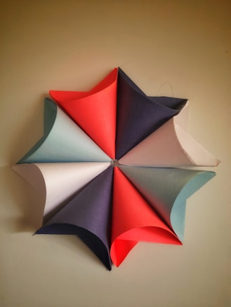 A paper craft piece forming a star-like pattern is displayed. Each section is composed of colored paper cones arranged in a symmetrical layout. The colors include red, blue, white, and light blue, creating a visually appealing geometric design.