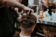 A barber is giving a haircut to a client. The focus is on the hands of the barber using scissors to trim the client's hair. The client has short, dark hair, and the setting appears to be a busy barbershop with various tools and products visible in the background.
