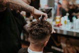 A barber is giving a haircut to a client. The focus is on the hands of the barber using scissors to trim the client's hair. The client has short, dark hair, and the setting appears to be a busy barbershop with various tools and products visible in the background.