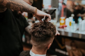 A barber is giving a haircut to a client. The focus is on the hands of the barber using scissors to trim the client's hair. The client has short, dark hair, and the setting appears to be a busy barbershop with various tools and products visible in the background.