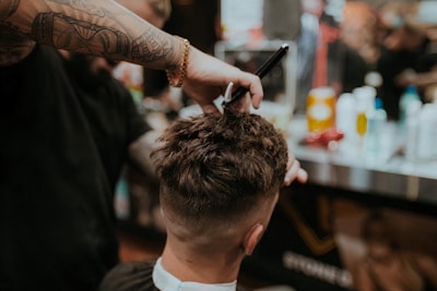 A barber is giving a haircut to a client. The focus is on the hands of the barber using scissors to trim the client's hair. The client has short, dark hair, and the setting appears to be a busy barbershop with various tools and products visible in the background.