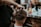 A barber is giving a haircut to a client. The focus is on the hands of the barber using scissors to trim the client's hair. The client has short, dark hair, and the setting appears to be a busy barbershop with various tools and products visible in the background.