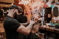A barber is giving a haircut to a client in a stylish and colorful barbershop. The barber, wearing a black cap and t-shirt, focuses intently as he uses scissors to trim the client's hair. The shop's walls are adorned with vibrant graffiti art and various decorations, adding to the lively atmosphere.