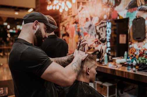 A barber is giving a haircut to a client in a stylish and colorful barbershop. The barber, wearing a black cap and t-shirt, focuses intently as he uses scissors to trim the client's hair. The shop's walls are adorned with vibrant graffiti art and various decorations, adding to the lively atmosphere.