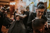 People are in a dimly lit barbershop, where multiple barbers are working on cutting client's hair. One barber in the foreground is focused on shaving or shaping a client's hair. The decor is warm and modern, with bright lights in the background providing a cozy atmosphere.