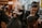 People are in a dimly lit barbershop, where multiple barbers are working on cutting client's hair. One barber in the foreground is focused on shaving or shaping a client's hair. The decor is warm and modern, with bright lights in the background providing a cozy atmosphere.