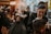 People are in a dimly lit barbershop, where multiple barbers are working on cutting client's hair. One barber in the foreground is focused on shaving or shaping a client's hair. The decor is warm and modern, with bright lights in the background providing a cozy atmosphere.