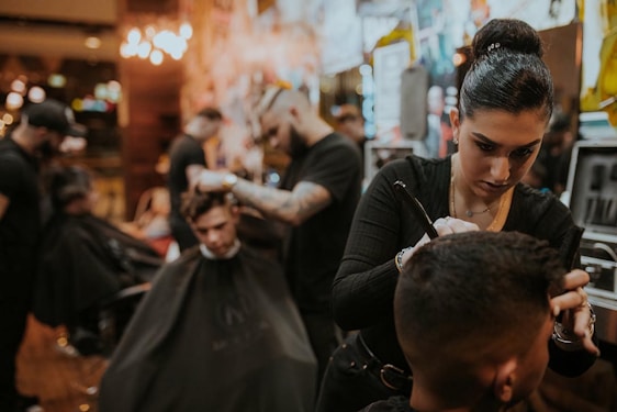 People are in a dimly lit barbershop, where multiple barbers are working on cutting client's hair. One barber in the foreground is focused on shaving or shaping a client's hair. The decor is warm and modern, with bright lights in the background providing a cozy atmosphere.