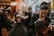 People are in a dimly lit barbershop, where multiple barbers are working on cutting client's hair. One barber in the foreground is focused on shaving or shaping a client's hair. The decor is warm and modern, with bright lights in the background providing a cozy atmosphere.