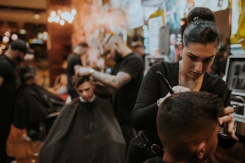 People are in a dimly lit barbershop, where multiple barbers are working on cutting client's hair. One barber in the foreground is focused on shaving or shaping a client's hair. The decor is warm and modern, with bright lights in the background providing a cozy atmosphere.