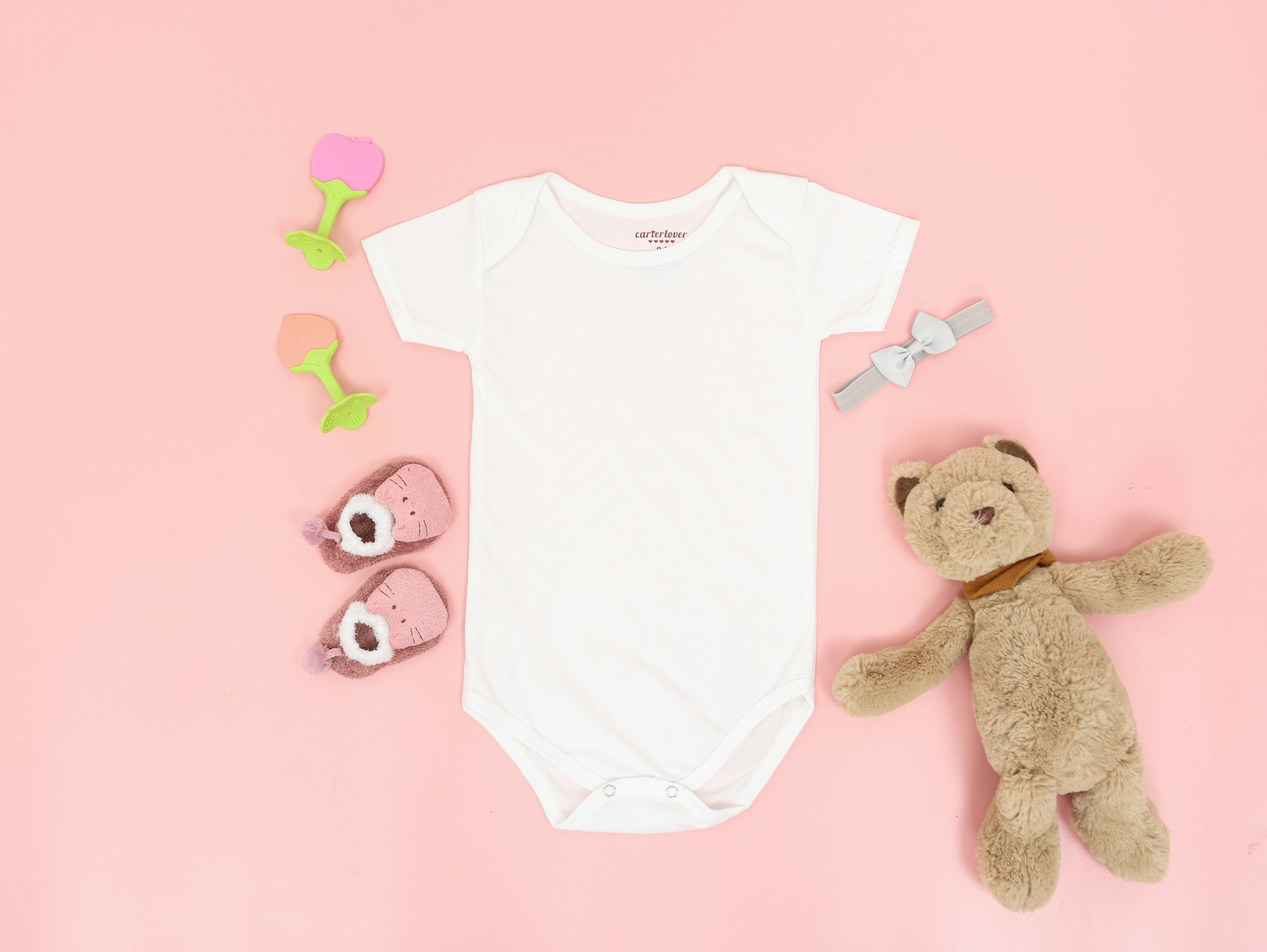 A onesie lying on a pink surface next to baby toys and a cute teddy bear.