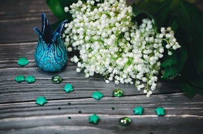 A beautifully crafted blue ceramic vase with intricate patterns sits on a rustic wooden surface. Beside it is a lush bouquet of delicate white flowers, likely lilies of the valley, with rich green leaves. Scattered around are teal origami stars and glass beads, enhancing the elegant composition with a whimsical touch.