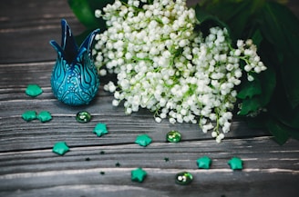 A beautifully crafted blue ceramic vase with intricate patterns sits on a rustic wooden surface. Beside it is a lush bouquet of delicate white flowers, likely lilies of the valley, with rich green leaves. Scattered around are teal origami stars and glass beads, enhancing the elegant composition with a whimsical touch.