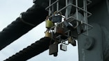A collection of various padlocks attached to a metal grate structure. The locks are of different sizes and colors, indicating a diverse range of origins. They hang from a lattice of metal bars situated against a background of thick, black cables.