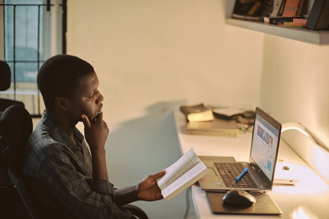 Young man reading a book with an open laptop in front of him