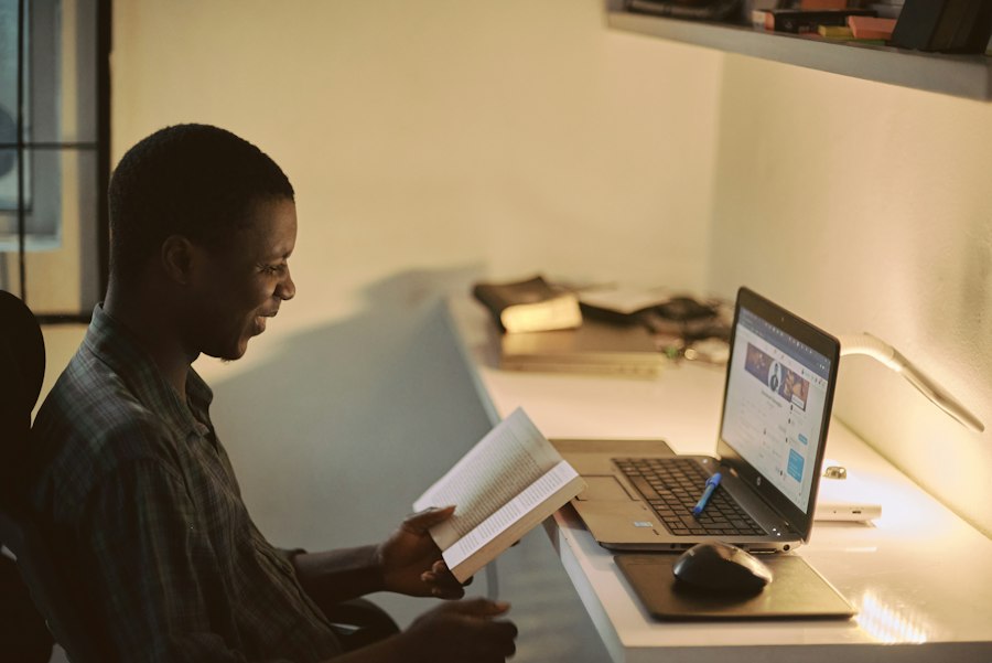 Man reading a book beside a laptop on a desk