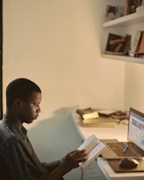 A person is sitting at a desk reading a book, with a laptop open in front of them. The desk is organized with some books and items on the shelves above. The lighting is warm and soft, creating a cozy atmosphere.