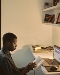 A person is sitting at a desk reading a book, with a laptop open in front of them. The desk is organized with some books and items on the shelves above. The lighting is warm and soft, creating a cozy atmosphere.