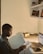 A person is sitting at a desk reading a book, with a laptop open in front of them. The desk is organized with some books and items on the shelves above. The lighting is warm and soft, creating a cozy atmosphere.