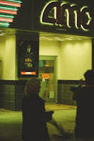 A dimly lit entrance to a movie theater with an AMC sign above. The area is bathed in a greenish glow from the neon lights. Two blurred figures are in the foreground, possibly walking past or standing near. A movie poster is visible on the wall beside the entrance.