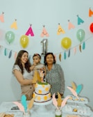 A birthday celebration scene with three people around a table. A woman is holding a young child, and another woman stands beside them. The table features a decorated two-tier cake with yellow flowers and a paper teepee decoration. Overhead, a garland of colorful balloons and paper cutouts forms the word “Pia” with a metallic number one balloon indicating it might be a first birthday. Decorative feathers and a lace tablecloth add to the festive atmosphere.