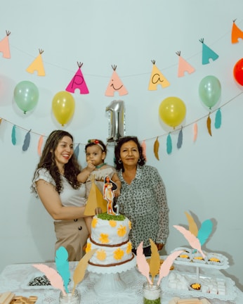 A birthday celebration scene with three people around a table. A woman is holding a young child, and another woman stands beside them. The table features a decorated two-tier cake with yellow flowers and a paper teepee decoration. Overhead, a garland of colorful balloons and paper cutouts forms the word “Pia” with a metallic number one balloon indicating it might be a first birthday. Decorative feathers and a lace tablecloth add to the festive atmosphere.