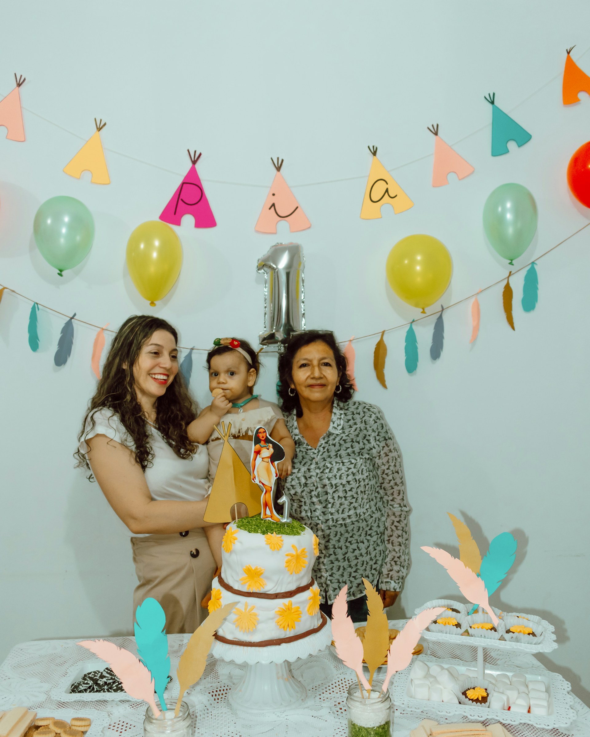 A birthday celebration scene with three people around a table. A woman is holding a young child, and another woman stands beside them. The table features a decorated two-tier cake with yellow flowers and a paper teepee decoration. Overhead, a garland of colorful balloons and paper cutouts forms the word “Pia” with a metallic number one balloon indicating it might be a first birthday. Decorative feathers and a lace tablecloth add to the festive atmosphere.